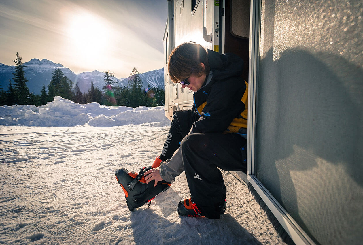 Man putting on ski boots while sitting on step of CanaDream winter RV